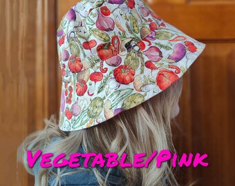 May include: A white bucket hat with a colorful vegetable print. The hat has a pink band and the text "VEGETABLE/PINK DAISY" is printed on the front.