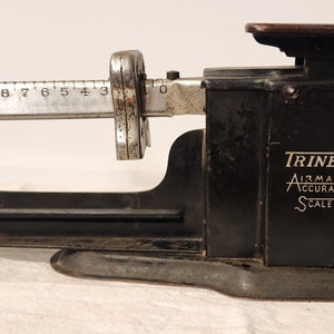 May include: A black Triner Airmail Accuracy Scale with a silver dial and a wooden platform. The scale is designed to weigh letters and packages.