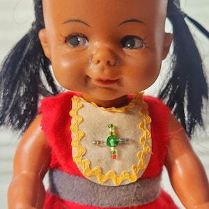 Könnte beinhalten: Eine Vintage-Puppe mit dunklen Haaren in Zöpfen, die ein rotes Kleid mit grünem und gelbem Saum trägt. Die Puppe hat eine Perlenkette mit einem grünen Stein.