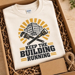 May include: A cream-colored crew neck t-shirt featuring a graphic of a building, a pipe wrench, and a flashlight in black and yellow. The text reads KEEP THE BUILDING RUNNING in bold black letters. The shirt is folded inside a cardboard gift box.