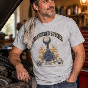 May include: A light grey short-sleeve t-shirt featuring a graphic of a manual car gear shifter. The text reads ENDANGERED SPECIES, MANUALIS TRANSMISSIONES, NATIVE HABITAT: THE LEFT LANE, and STATUS: CRITICALLY RARE, with a 6-speed shift pattern.