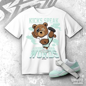 Puede incluir: Camiseta blanca con un oso de dibujos animados que lleva una camiseta blanca y pantalones cortos azul claro, levantando una pierna. El texto de la camiseta dice "KICKS SPEAK LOUDER THAN WORDS". Un par de zapatillas blancas y azul claro están en primer plano.