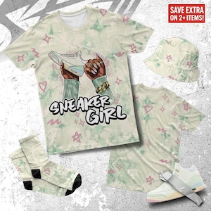 Könnte beinhalten: Ein cremefarbenes T-Shirt, ein Fischerhut und ein Socken-Set mit einem sich wiederholenden Muster aus rosa Sternen und Herzen. Das T-Shirt zeigt den Text "SNEAKER GIRL" und eine Illustration von Händen, die einen Sneaker halten. Ein passender Sneaker ist ebenfalls abgebildet.