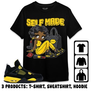 May include: Black t-shirt with a cartoon illustration of a Black woman wearing yellow clothing and holding a purse. The text "SELF MADE" is printed in yellow letters on the shirt. The image also includes a pair of black and yellow sneakers.