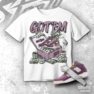 Könnte beinhalten: Ein weißes T-Shirt mit einer grafischen Illustration eines lila Schuhkartons, der mit Bargeld und einem Paar lila Sneakern gefüllt ist. Der Text GOT'EM ist in Lila über dem Karton gedruckt, und die Zahl 23 befindet sich an der Seite. Ein einzelner lila Sneaker ist unten rechts abgebildet.