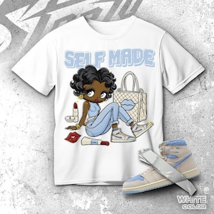 Puede incluir: Camiseta blanca con una ilustración de dibujos animados de una mujer con vaqueros azules y zapatillas deportivas, con el texto "SELF MADE" en azul claro. La ilustración incluye un bolso, pintalabios y maquillaje. Un par de zapatillas deportivas azules y beige también están en la imagen.