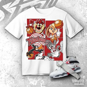 May include: White t-shirt featuring a red and white graphic of the Tune Squad basketball team with Taz, Lola Bunny, Bugs Bunny, and Daffy Duck. A pair of white and gray sneakers with red accents are in the foreground.