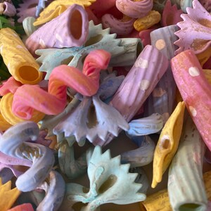 Pu&ograve; includere: Primo piano di varie forme di pasta dai colori pastello. L'immagine presenta farfalle, penne e altri tipi di pasta in tonalit&agrave; rosa, blu, giallo e viola. La pasta &egrave; disposta in primo piano, riempiendo l'inquadratura.
