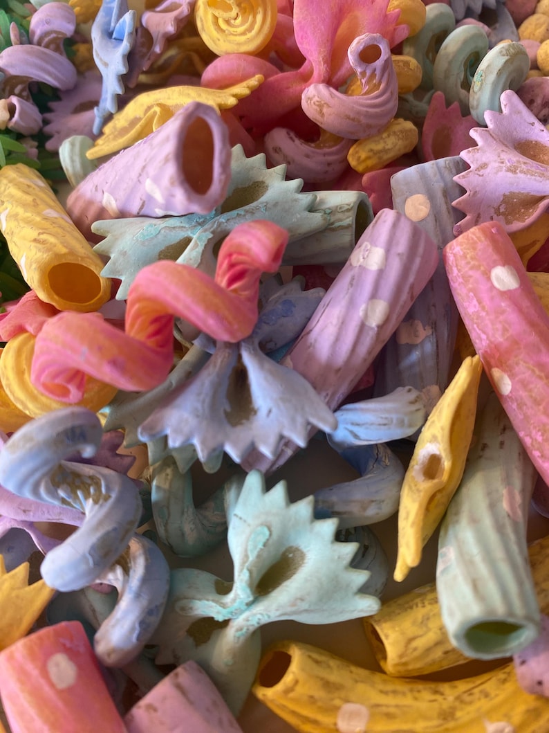 Pu&ograve; includere: Primo piano di varie forme di pasta dai colori pastello. L'immagine presenta farfalle, penne e altri tipi di pasta in tonalit&agrave; rosa, blu, giallo e viola. La pasta &egrave; disposta in primo piano, riempiendo l'inquadratura.