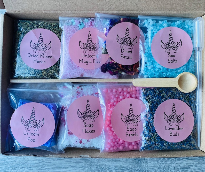 May include: A box set containing various bath and spa products. The set includes bags of dried herbs, bath salts, soap flakes, dried petals, sea salts, sago pearls, and lavender buds. Each bag has a pink label with a unicorn design and product name.