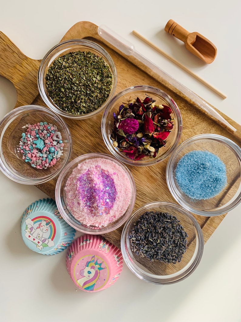 May include: A set of six small glass bowls filled with various ingredients, including dried herbs, flower petals, sprinkles, sugar, and lavender. Two unicorn-themed cupcake liners are also included.