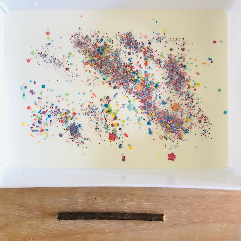 Pu&ograve; includere: Un vassoio rettangolare bianco riempito con un liquido cremoso e spruzzi colorati. Gli spruzzi sono un mix di forme e colori, tra cui stelle, cerchi e bastoncini. Un bastoncino marrone scuro poggia su una superficie di legno sottostante.
