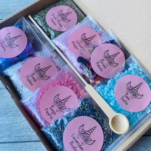 May include: A gift box filled with various bath products, including dried herbs, bath salts, and soap flakes. Each item is packaged in a clear bag and labeled with a pink sticker featuring a unicorn design. A small wooden scoop is included.