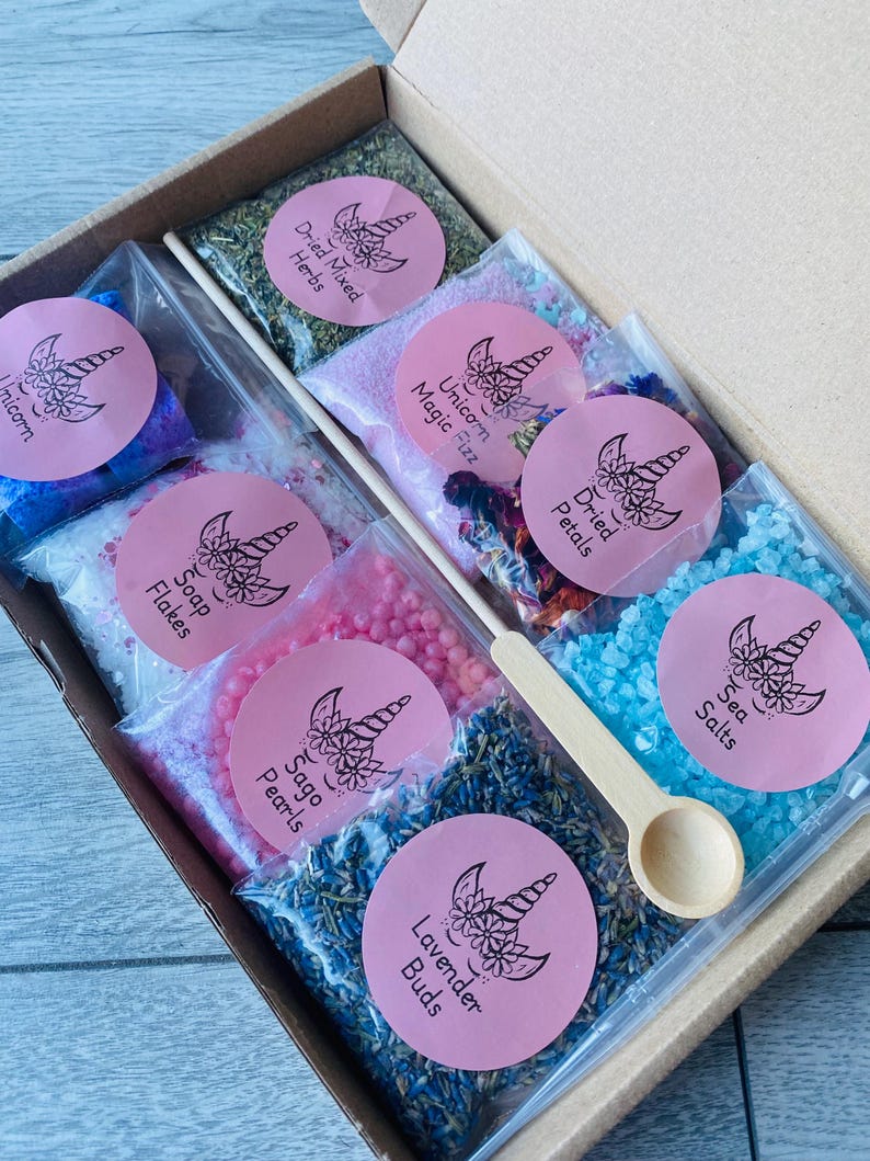 May include: A gift box filled with various bath products, including dried herbs, bath salts, and soap flakes. Each item is packaged in a clear bag and labeled with a pink sticker featuring a unicorn design. A small wooden scoop is included.