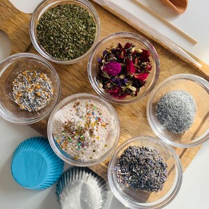 May include: A wooden tray with six small glass bowls filled with various ingredients, including dried herbs, flower petals, glitter, and sprinkles. Two blue and white cupcake liners are also included.