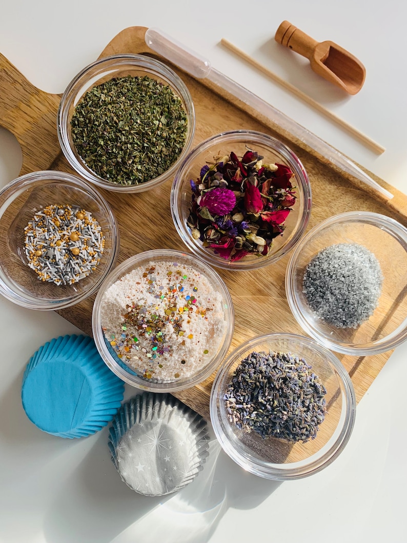 May include: A wooden tray with six small glass bowls filled with various ingredients, including dried herbs, flower petals, glitter, and sprinkles. Two blue and white cupcake liners are also included.