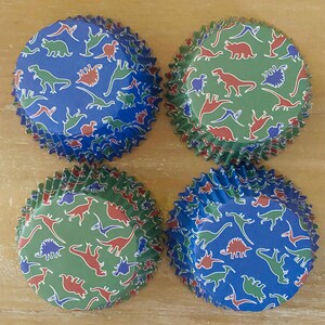 May include: Four cupcake liners with a dinosaur pattern. The liners are blue and green with red and white dinosaurs.