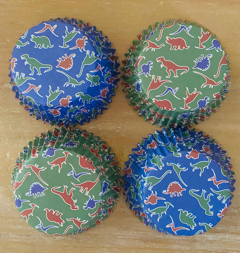 May include: Four cupcake liners with a dinosaur pattern. The liners are blue and green with red and white dinosaurs.