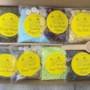 Könnte beinhalten: Eine Box mit acht kleinen Beuteln mit Badeprodukten, darunter getrocknete Kräuter, Sommer-Zauber-Fizz, getrocknete Blütenblätter, Meersalz, Strandsteine, Seifenflocken, Sagoperlen und Lavendelknospen. Jeder Beutel hat ein gelbes Etikett mit einer Strandillustration.