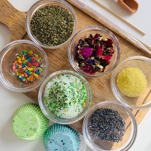 May include: A wooden board with six small glass bowls filled with various ingredients, including dried herbs, flower petals, sprinkles, green glitter, sugar, and lavender. Two green and blue cupcake liners with dinosaur designs are also on the board.