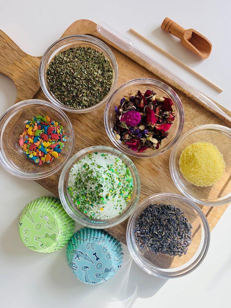 May include: A wooden board with six small glass bowls filled with various ingredients, including dried herbs, flower petals, sprinkles, green glitter, sugar, and lavender. Two green and blue cupcake liners with dinosaur designs are also on the board.