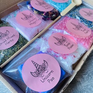 May include: A gift box filled with various bath products, including bath salts, dried herbs, and soap flakes. Each item is packaged in a clear plastic bag and labeled with a pink sticker featuring a unicorn design. A small wooden spoon and a wooden stick are included.