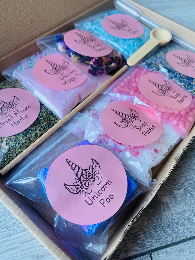 May include: A gift box filled with various bath products, including bath salts, dried herbs, and soap flakes. Each item is packaged in a clear plastic bag and labeled with a pink sticker featuring a unicorn design. A small wooden spoon and a wooden stick are included.