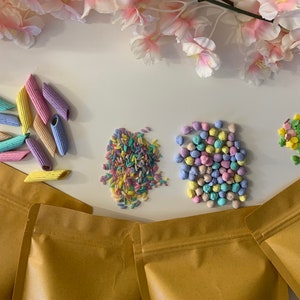 Pu&ograve; includere: Una vista dall'alto presenta pasta colorata, zuccherini e caramelle in tonalit&agrave; pastello. La pasta &egrave; a forma di penne e tinta in blu, giallo, rosa e viola. Zuccherini e caramelle sono anch'essi in colori pastello. Sacchetti di carta marrone sono in basso.