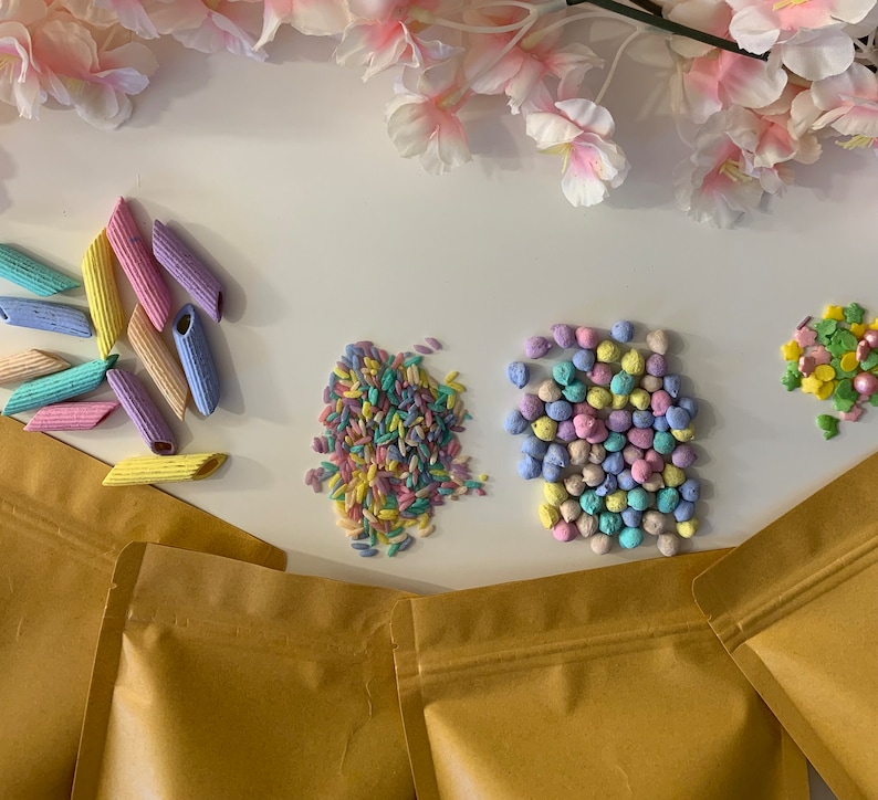 Pu&ograve; includere: Una vista dall'alto presenta pasta colorata, zuccherini e caramelle in tonalit&agrave; pastello. La pasta &egrave; a forma di penne e tinta in blu, giallo, rosa e viola. Zuccherini e caramelle sono anch'essi in colori pastello. Sacchetti di carta marrone sono in basso.