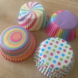 May include: Four colorful cupcake liners with different patterns. The liners are rainbow striped, purple with rainbow stripes, rainbow circles, and white with pink, blue, and green polka dots.