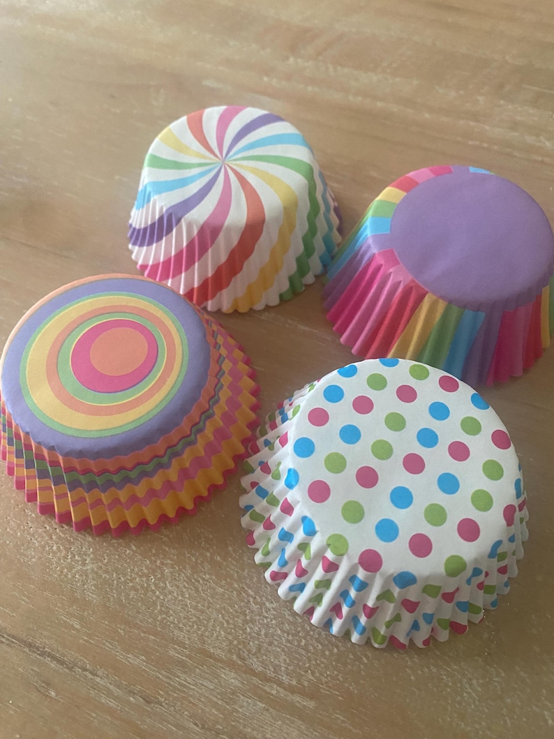 May include: Four colorful cupcake liners with different patterns. The liners are rainbow striped, purple with rainbow stripes, rainbow circles, and white with pink, blue, and green polka dots.