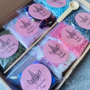 May include: A box set containing various bath products. The set includes small bags of blue sea salts, dried petals, lavender, sage pearls, soap flakes, and dried mixed herbs. Each bag has a pink label with a unicorn design. A small wooden spoon and a dropper are included.
