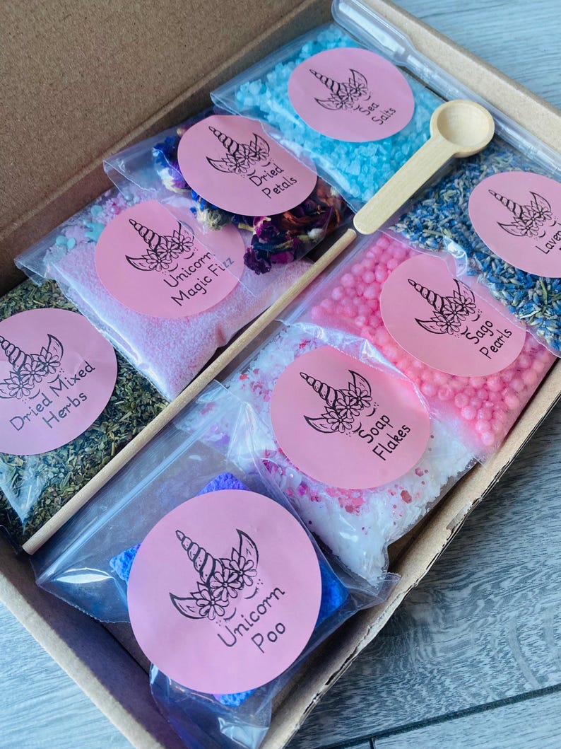 May include: A box set containing various bath products. The set includes small bags of blue sea salts, dried petals, lavender, sage pearls, soap flakes, and dried mixed herbs. Each bag has a pink label with a unicorn design. A small wooden spoon and a dropper are included.