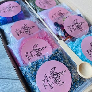 May include: A gift box filled with various bath products, including lavender buds, soap flakes, and dried petals. Each product is packaged in a clear bag with a pink label featuring a unicorn design. A wooden spoon is included.