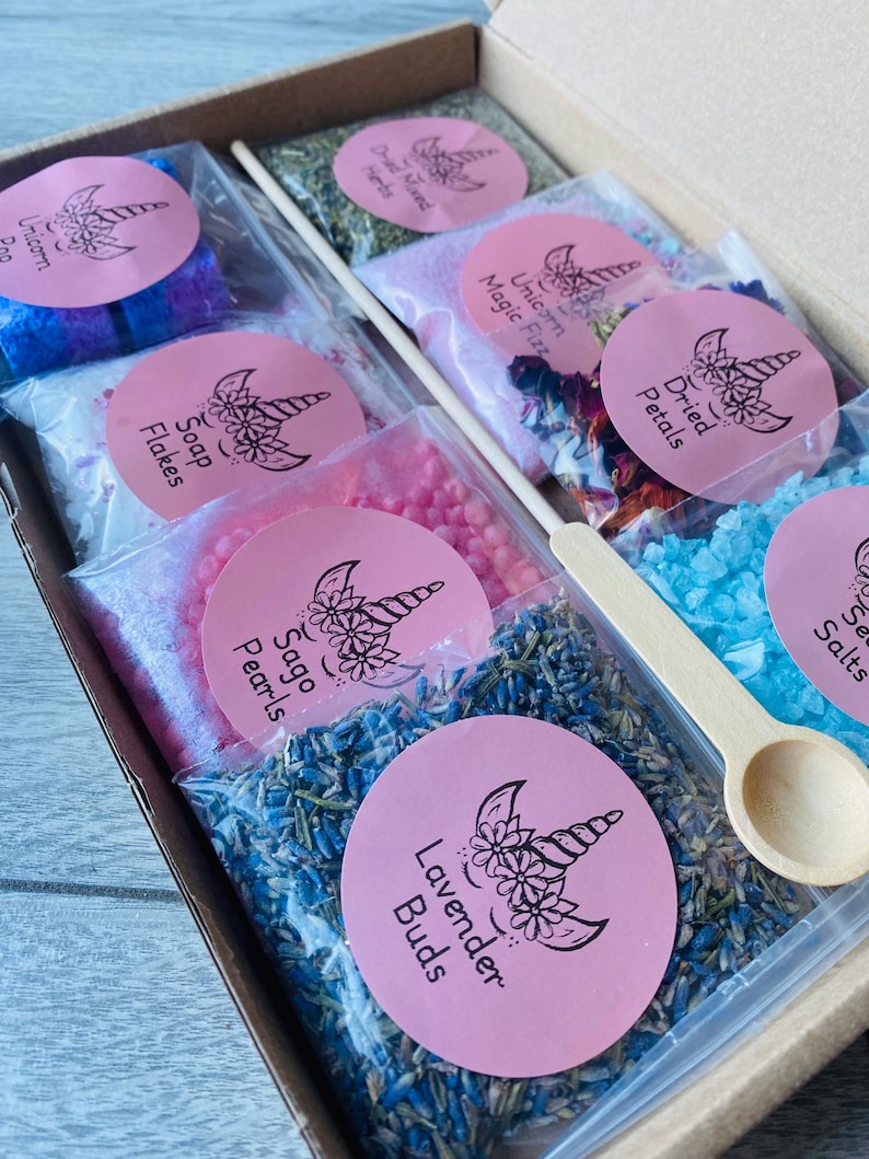May include: A gift box filled with various bath products, including lavender buds, soap flakes, and dried petals. Each product is packaged in a clear bag with a pink label featuring a unicorn design. A wooden spoon is included.