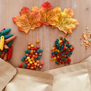 Puede incluir: Una colección de artículos sensoriales de temática otoñal, que incluyen pasta de colores, espolvorear de colores otoñales y pequeñas bolas de colores. Los artículos están dispuestos sobre una superficie de madera con bolsas de papel marrón en el fondo.