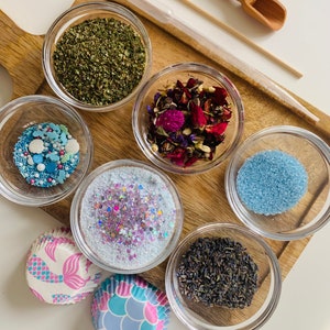 May include: Six small glass bowls filled with various ingredients, including dried herbs, flower petals, glitter, and sprinkles. Two mermaid-themed cupcake liners are also included.