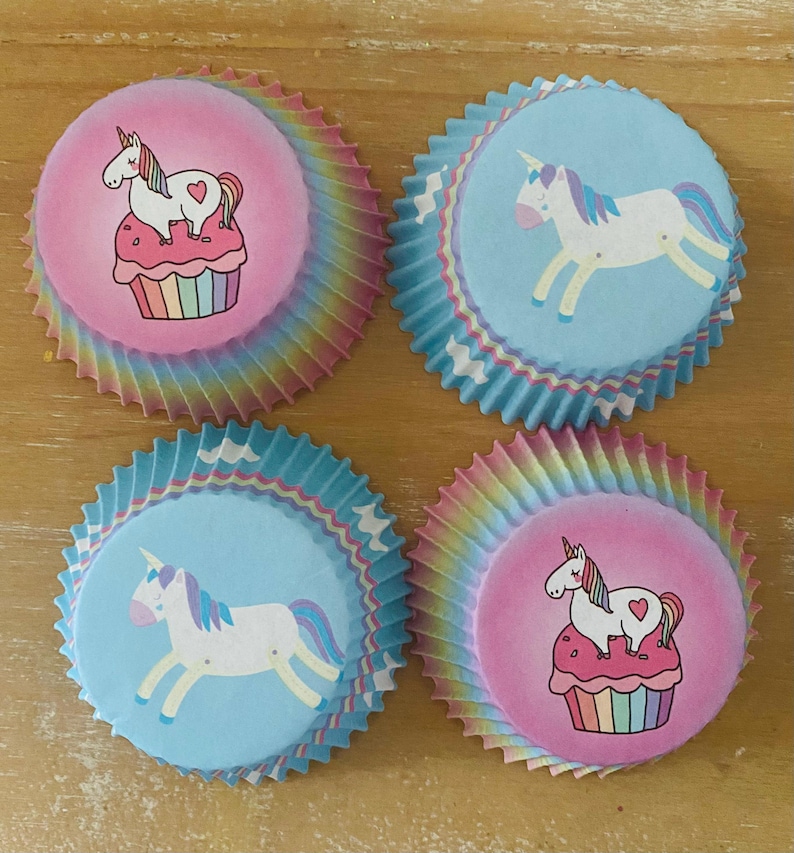May include: Four cupcake liners with unicorn designs. Two liners feature a unicorn on a cupcake, set against a pink background. The other two have a unicorn on a blue background. The liners have a rainbow-colored, ruffled edge.