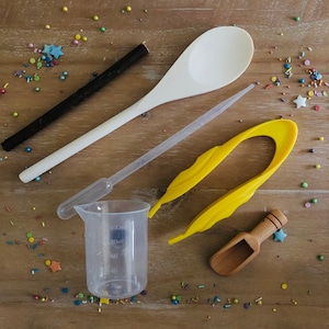 Pu&ograve; includere: Una collezione di strumenti per la cottura, tra cui un cucchiaio di legno bianco, un misurino di plastica trasparente, una pinzetta di plastica gialla, un piccolo misurino di legno, un bastoncino nero e un contagocce di plastica trasparente.  Sono sparsi confettini colorati.
