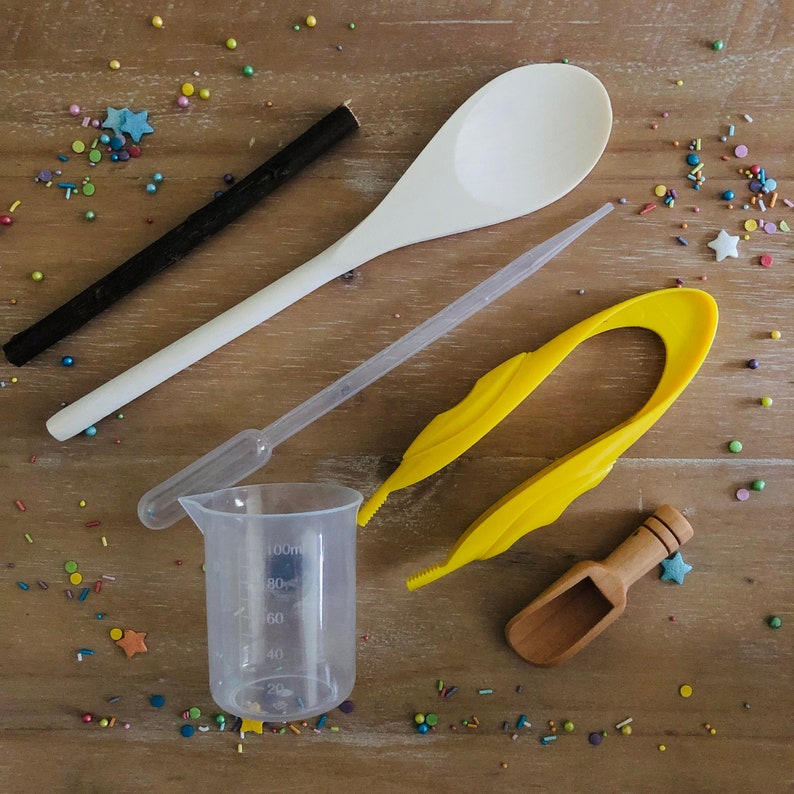 Pu&ograve; includere: Una collezione di strumenti per la cottura, tra cui un cucchiaio di legno bianco, un misurino di plastica trasparente, una pinzetta di plastica gialla, un piccolo misurino di legno, un bastoncino nero e un contagocce di plastica trasparente.  Sono sparsi confettini colorati.