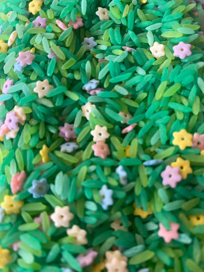 Pu&ograve; includere: Primo piano di un riempitivo per contenitore sensoriale con riso verde e zuccherini a forma di stella colorati. Il riso &egrave; tinto in varie tonalit&agrave; di verde, che assomigliano all'erba. Gli zuccherini sono di colori pastello, tra cui giallo, rosa e azzurro.