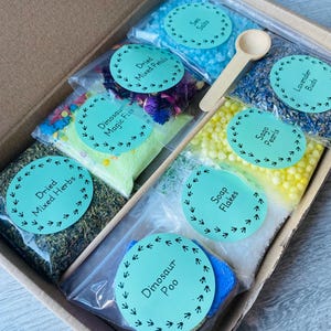 May include: A box filled with small bags of bath products. The bags are labeled with different names, including "Dried Mixed Herbs", "Dinosaur Magic Fizz", "Dried Mixed Petals", "Sea Salts", "Soap Flakes", "Sago Pearls", "Dinosaur Poo", and "Lavender Buds".