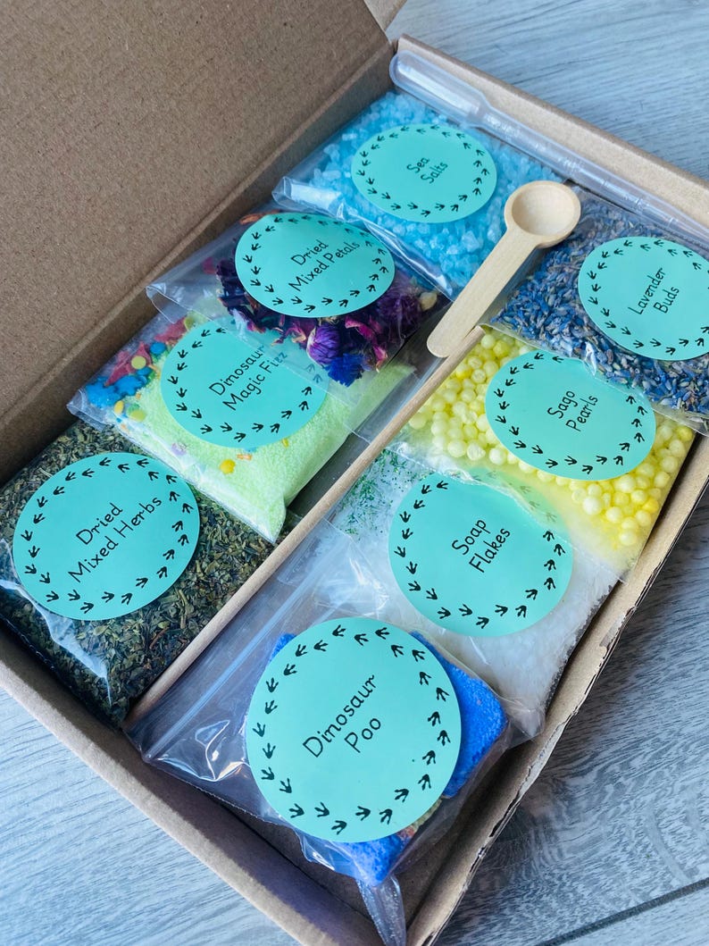 May include: A box filled with small bags of bath products. The bags are labeled with different names, including "Dried Mixed Herbs", "Dinosaur Magic Fizz", "Dried Mixed Petals", "Sea Salts", "Soap Flakes", "Sago Pearls", "Dinosaur Poo", and "Lavender Buds".
