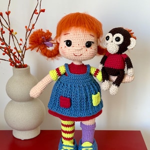 Könnte beinhalten: Eine handgefertigte Häkelpuppe mit leuchtend orangefarbenem Haar in Zöpfen, die ein blaues Trägerkleid über einem roten Oberteil mit gelben Ärmeln trägt. Die Puppe hat ungleiche Socken, eine gelb-rot gestreift und eine lila, und hält einen kleinen Affen.