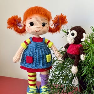 May include: A crocheted doll with bright orange hair in pigtails, wearing a blue pinafore dress over a red shirt, yellow and red striped stockings, one purple sock, and blue shoes, accompanied by a small brown and cream crocheted monkey toy.