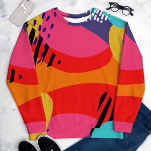 Puede incluir: Una sudadera de cuello redondo con un diseño abstracto vibrante. El patrón presenta formas grandes y superpuestas en tonos de rojo, naranja, rosa, amarillo, morado y turquesa, acentuadas con líneas negras y detalles blancos. La sudadera tiene mangas largas.