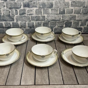 Puede incluir: Seis tazas de té blancas y platillos con un borde decorativo dorado y verde están dispuestas sobre una superficie de madera. Las tazas de té tienen un diseño clásico, perfecto para servir té o café.