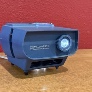 May include: A blue View-Master Entertainer Projector, a vintage device with a lens and a dial. The device has the text "View-Master Entertainer Projector" and the "gaf" logo. The projector is on a wooden surface.