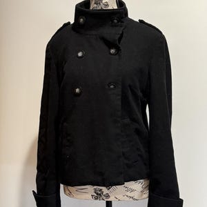 May include: A black double-breasted jacket with a high collar and button details. The jacket has a tailored fit and features buttoned cuffs and shoulder epaulets. The jacket is displayed on a mannequin.