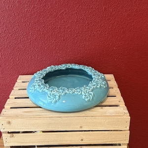 May include: A round, turquoise ceramic planter with a decorative floral border. The planter sits on a wooden pallet against a red wall. The planter is a vibrant shade of blue.